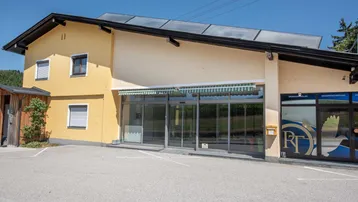 Gelbes Gebäude mit großen Glasfronten, Vordach und Solaranlage auf dem Dach, umgeben von Parkplätzen.