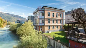 Historisches Gebäude am Flussufer mit grüner Umgebung und Bergen im Hintergrund.