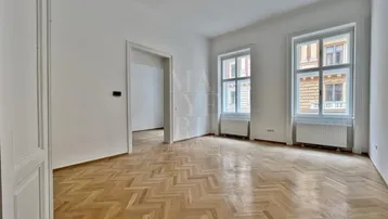 Helles Zimmer mit Fischgrätparkettboden, hohen Decken und großen Fenstern, das viel Tageslicht hereinlässt.