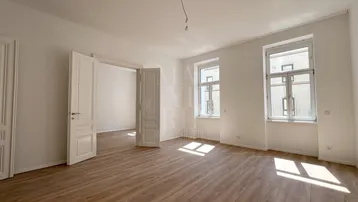 Helles, leeres Wohnzimmer mit Holzboden und großen Fenstern, das viel Tageslicht hereinlässt.