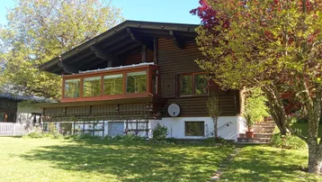 Charmantes Holzhaus mit gepflegtem Rasen, großen Bäumen und verglastem Wintergarten unter blauem Himmel.