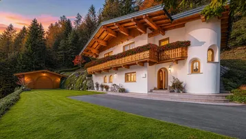 Traditionelles Alpenchalet mit weißer Fassade, Holzbalkonen und gepflegtem Vorgarten bei Sonnenuntergang.