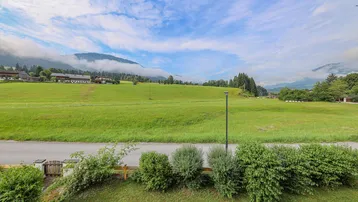 Weitläufiger Blick über grüne Wiesen und sanfte Hügel mit Bergen im Hintergrund unter bewölktem Himmel.