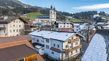 Luftaufnahme eines verschneiten Dorfes mit Kirche und Bergen im Hintergrund.