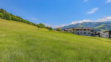 Weitläufige grüne Wiese mit Blick auf die umliegende Berglandschaft und entfernte Gebäude.