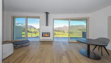 Helles Wohnzimmer mit Kamin, Holzboden und großen Schiebetüren, die einen weiten Blick auf die Berge und grüne Landschaft bieten.