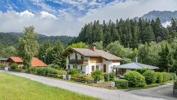 Traditionelles Haus im alpenländischen Stil mit Holzbalkonen und gepflegtem Garten, umgeben von grüner Natur und Bergen.