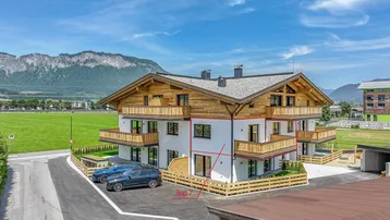 Modernes Mehrfamilienhaus mit Holzfassade und Balkonen, eingebettet in eine grüne Landschaft mit Bergblick.