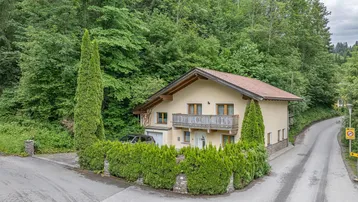 Freistehendes Haus mit Satteldach, Balkon und gepflegtem Vorgarten, umgeben von viel Grün.