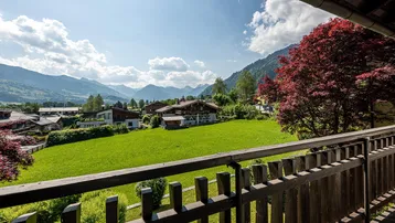 Panoramablick vom Balkon über grüne Wiesen, Häuser und die umliegende Berglandschaft an einem sonnigen Tag.