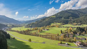 Panoramablick auf ein grünes Tal mit Bergen, einer Straße und verstreuten Häusern unter blauem Himmel.