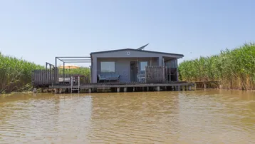 Kleines Haus auf Stelzen über dem Wasser, umgeben von Schilf, unter blauem Himmel.