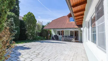Große gepflasterte Terrasse mit Gartenmöbeln und Grillbereich, umgeben von viel Grün.
