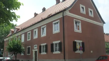 Rötlich-braunes Mehrfamilienhaus mit Satteldach, weißen Fenstern und dunklen Fensterläden an einer Straße.