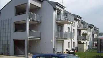 Mehrfamilienhaus mit moderner Fassade, Balkonen und Außenparkplätzen unter bewölktem Himmel.
