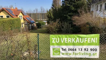 Blick auf das Grundstück mit einem 'Zu Verkaufen'-Schild am Zaun und einem Haus im Hintergrund.
