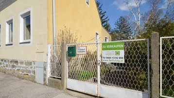 Gelbe Hausfassade mit einem Gartentor und einem "Zu Verkaufen"-Schild unter blauem Himmel.
