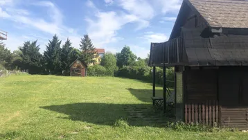 Weitläufige Rasenfläche mit einem kleinen Holzhaus und Bäumen unter blauem Himmel.