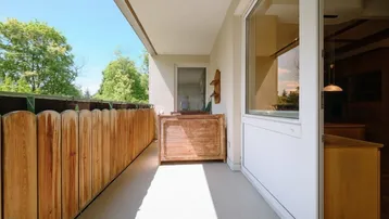 Langer, schmaler Balkon mit Holzzaun und Stauraum, umgeben von grünen Bäumen.