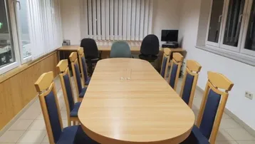 Konferenzraum mit großem Holztisch und blauen Stühlen, umgeben von Fenstern mit Jalousien.