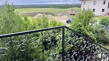Weitblick über die Landschaft mit Bäumen und einem Haus, gesehen von einem Balkon mit schmiedeeisernem Geländer.