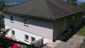 Das Haus präsentiert sich mit einer hellen Fassade und einem dunklen Dach, umgeben von einer malerischen Berglandschaft.