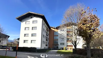 Mehrfamilienhaus mit weißer Fassade und Bäumen im Vordergrund unter blauem Himmel.