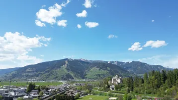Weitläufige Landschaftsaufnahme mit Bergen, Stadt, Fußballfeld und einem Schloss in der Ferne.