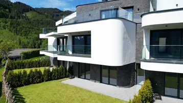 Moderne Architektur mit mehreren Balkonen, umgeben von grünen Gärten und Bergen im Hintergrund.