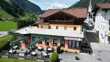 Luftaufnahme des Gebäudes mit Restaurant, Außenterrasse und umliegender Berglandschaft in einem alpinen Dorf.