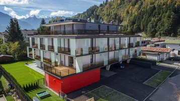 Modernes Mehrfamilienhaus mit Balkonen und Bergblick, umgeben von grüner Natur und Bäumen.
