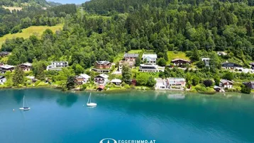 Panoramablick auf den See mit Häusern am Ufer und bewaldeten Bergen im Hintergrund unter blauem Himmel.