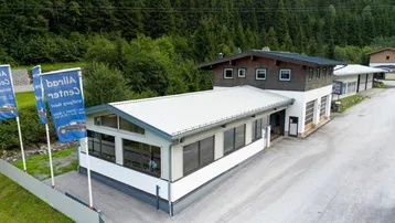 Luftaufnahme eines Gewerbegebäudes mit großen Fenstern und einem bewaldeten Berghang im Hintergrund.