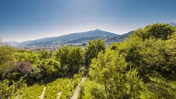 Panoramablick auf die Stadt im Tal, umgeben von grünen Hügeln und schneebedeckten Bergen.