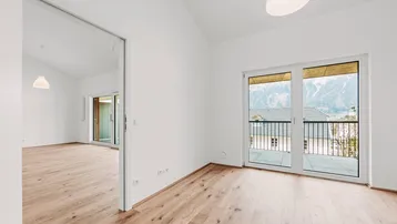 Helles, leeres Zimmer mit Holzboden, großen Fenstern und Zugang zu einem Balkon mit Bergblick.