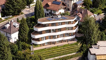 Moderne Wohnanlage mit geschwungenen Balkonen und Solarpaneelen auf dem Dach, umgeben von Bäumen.