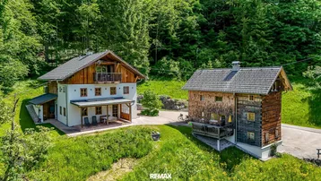 Zwei traditionelle Häuser in alpiner Umgebung, umgeben von grünen Wiesen und dichtem Wald.