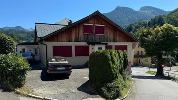 Traditionelles Haus mit Holzelementen und roten Fensterläden, umgeben von Bergen und Grünflächen.