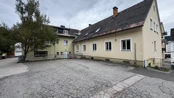 Außenansicht eines Gebäudes mit großem gepflasterten Vorplatz und einem Baum.