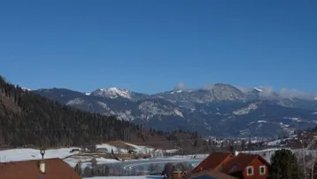 Panoramablick auf eine verschneite Winterlandschaft mit majestätischen Bergen und vereinzelten Häusern unter blauem Himmel.