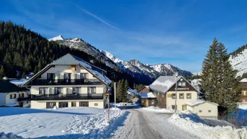 Winterliche Berglandschaft mit einem Mehrfamilienhaus und verschneiten Gipfeln unter blauem Himmel.