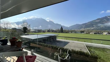 Geräumiger Balkon mit Holzboden, Glasgeländer und weitem Blick auf die Berge und grüne Landschaft.