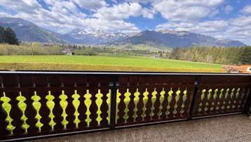 Blick vom Balkon über grüne Felder auf die beeindruckende Bergkulisse unter blauem Himmel.