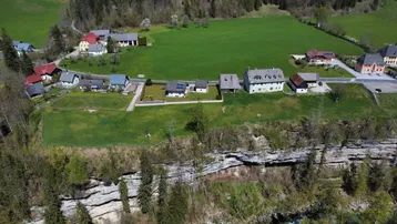 Luftaufnahme einer ländlichen Siedlung mit mehreren Häusern, grünen Wiesen und einem Fluss am Fuße einer Klippe.
