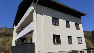 Mehrstöckiges Gebäude mit Balkonen und weißer Fassade vor einer grünen Hügellandschaft.