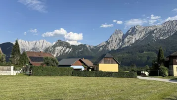 Weitläufige Wiesenfläche mit Blick auf traditionelle Häuser und majestätische Berglandschaft unter blauem Himmel.