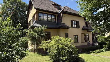 Charmantes Haus mit traditioneller Architektur, umgeben von üppigem Grün und altem Baumbestand.