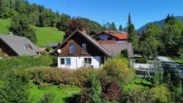 Haus in ländlicher Umgebung mit Satteldach und Holzbalkon, umgeben von viel Grün und Bergen.