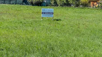 Grünes Grundstück mit einem 'Zu Verkaufen'-Schild, umgeben von Bäumen und Wohnhäusern.