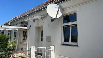 Weißes Einfamilienhaus mit Satellitenschüssel und kleinem Vorgarten unter blauem Himmel.